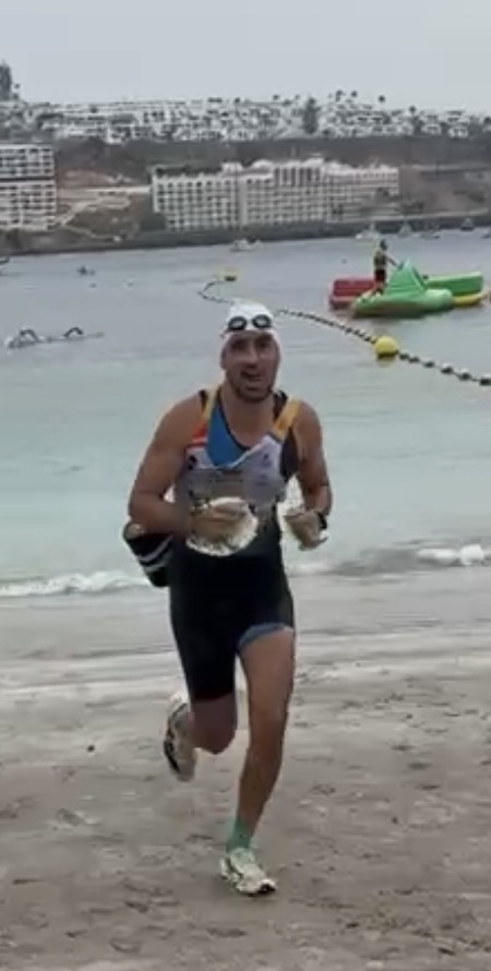 Alexis Garcia coming out of the water during a swimrun race, wetsuit half down, goggles on his head, with a bright coastline behind him.
