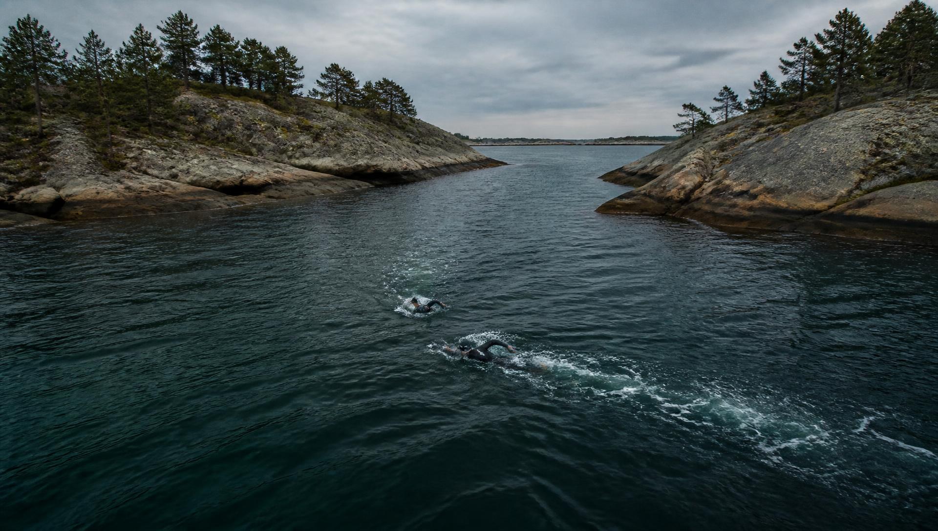 Two swimrun athletes crossing dark water between rocky islands